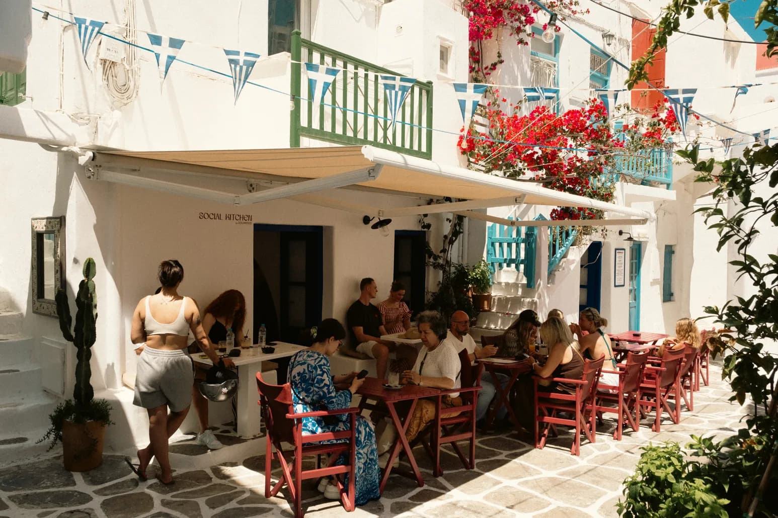 Guests relax at red wooden tables outside the whitewashed café in a Mykonos alley strung with Greek-flag bunting and red bougainvillea.