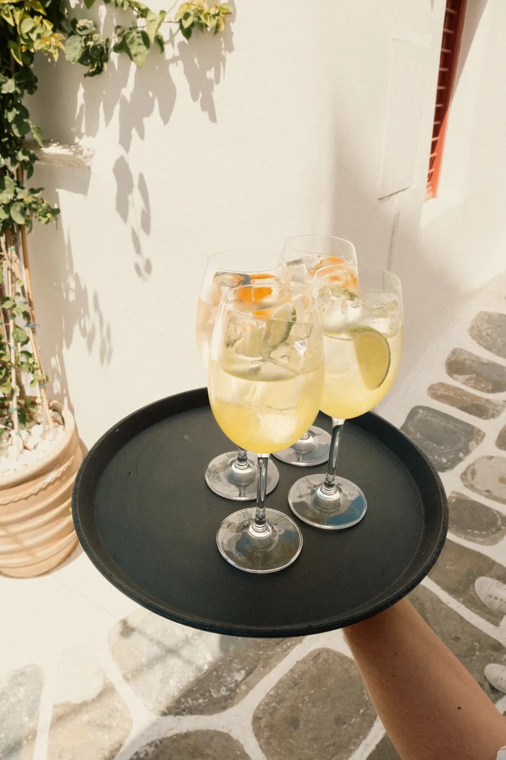 A server's tray holds four spritz glasses with citrus and lime, backlit against a whitewashed wall and stone-paved lane.