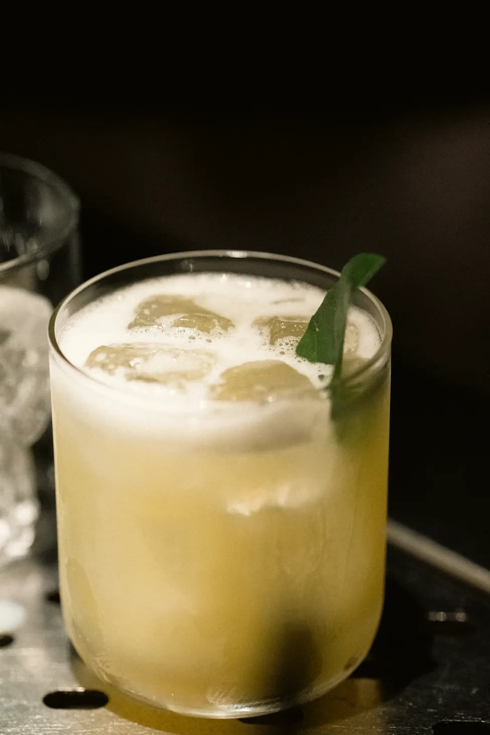 Close-up of a finished yellow sour cocktail with a thick foam cap and a single green leaf garnish.