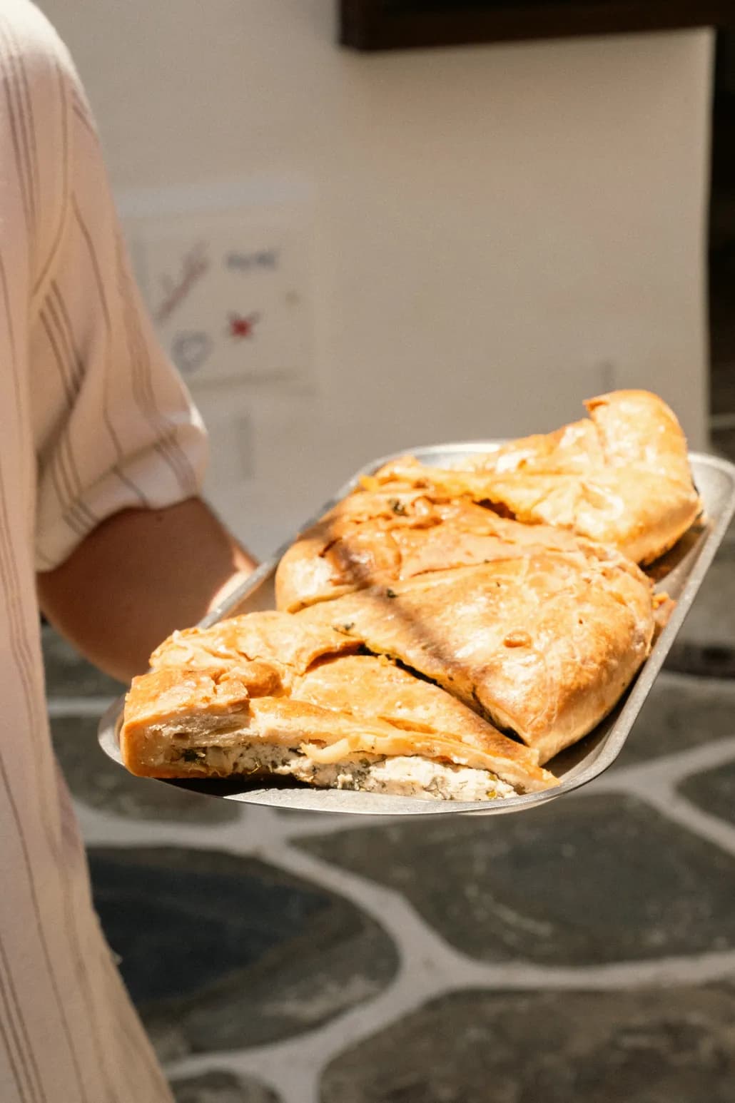 A staff member carries a metal tray of freshly baked savory pies through a sunlit whitewashed alley.