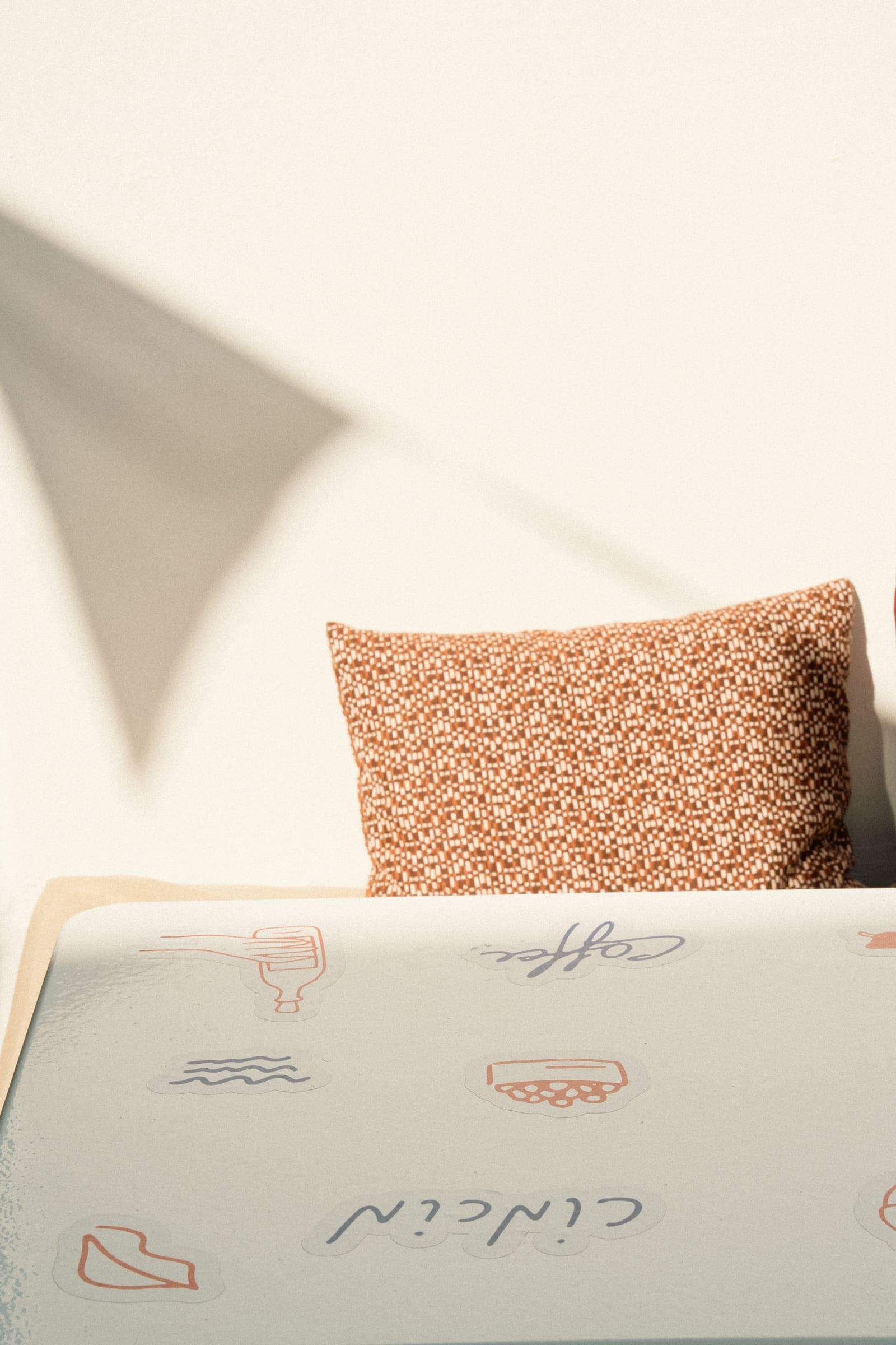 Rust-toned patterned cushion against a sunlit whitewashed wall with a sharp triangular shadow, above a pale tabletop scattered with playful illustrated stickers reading 'coffee' and 'cin cin'.