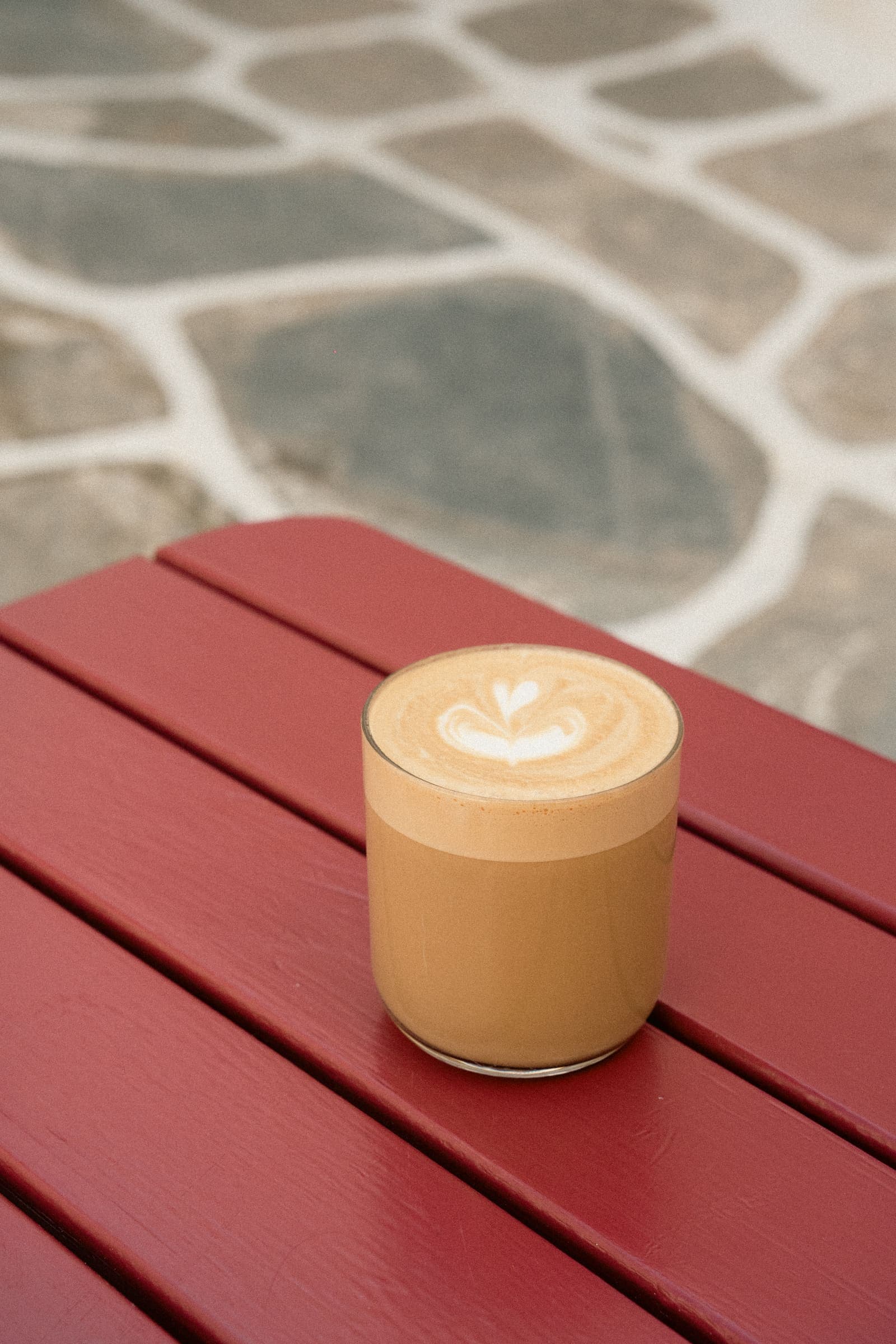 Flat white in a clear glass with heart-shaped latte art, set on a deep-red slatted café table above the grey painted flagstones of a Mykonos lane.