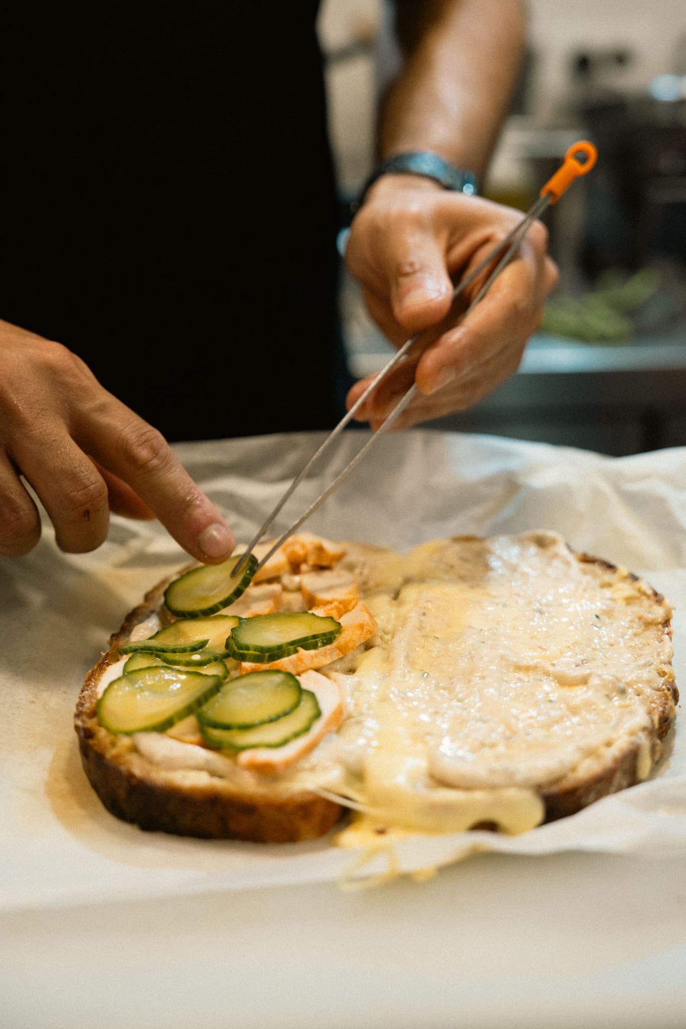 Hands with tweezers layer pickled cucumber slices onto an open sourdough sandwich oozing with melted cheese on parchment paper.