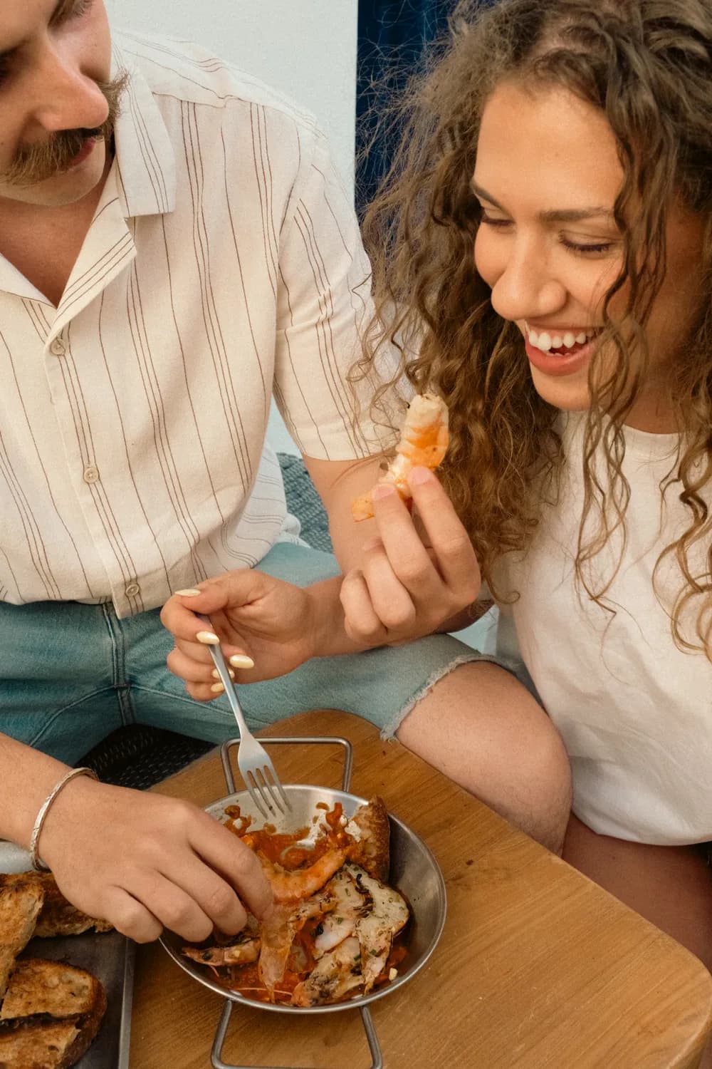 A laughing woman with curly hair holds a peeled prawn while sharing a pan of meze with a friend.