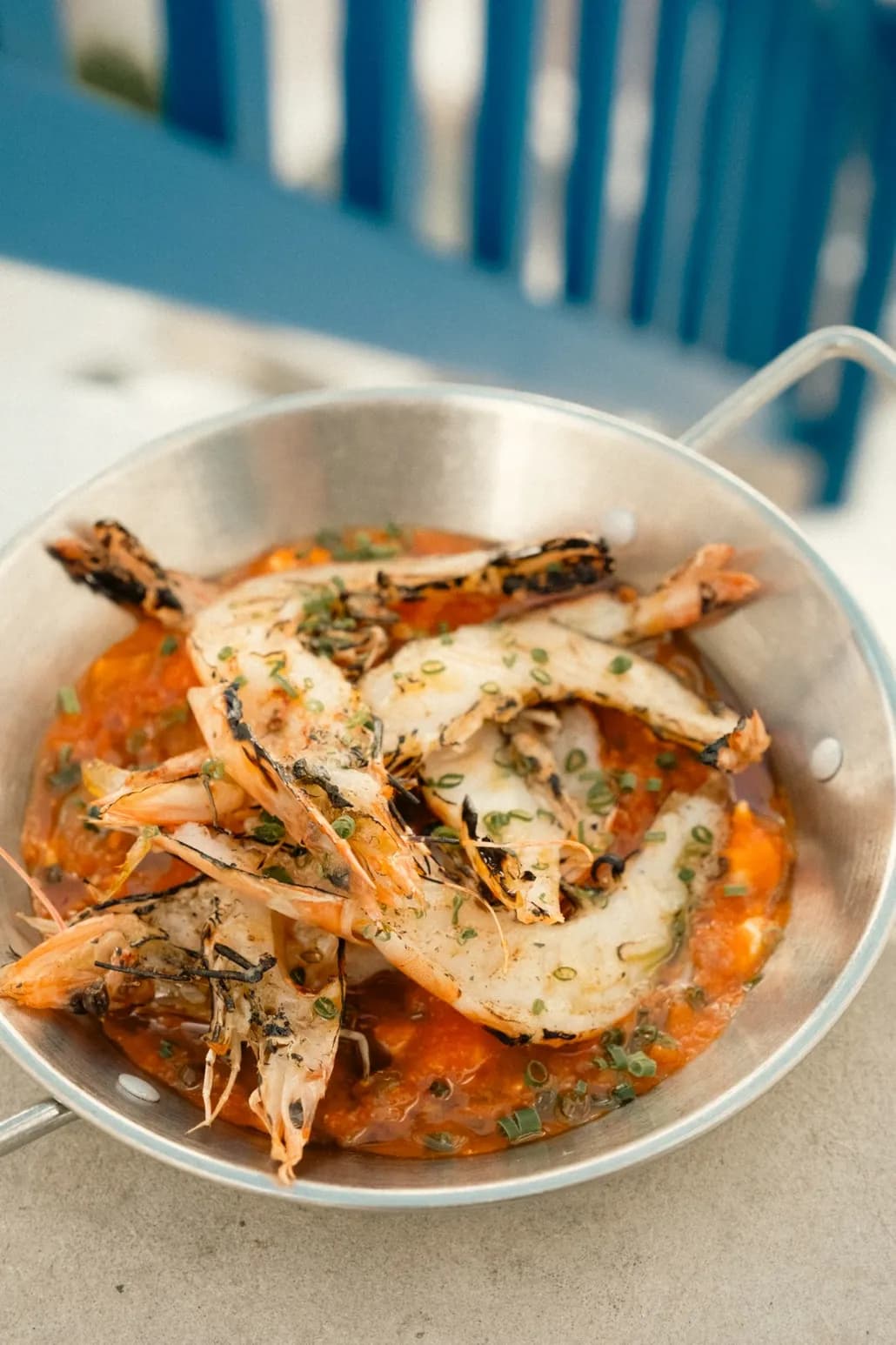 Grilled prawns piled in a steel pan of rich tomato sauce with fresh chives, on a whitewashed table beside a blue railing.